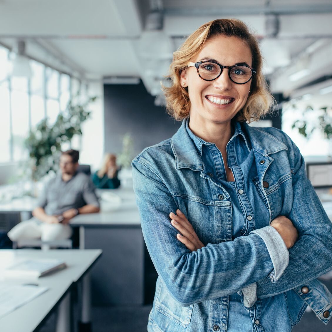 Successful creative female standing in office with colleagues in background. Happy woman executive standing in office with arms crossed.