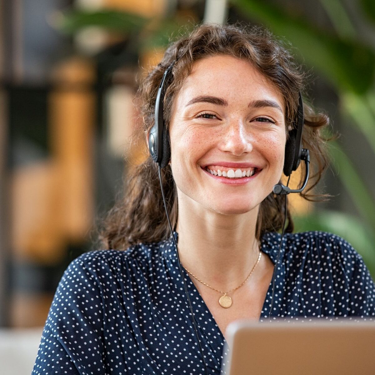 Portrait of smiling casual businesswoman working in a creative office. Smiling friendly woman working as call center agent for online support. Young woman in video call with headphones looking at camera.